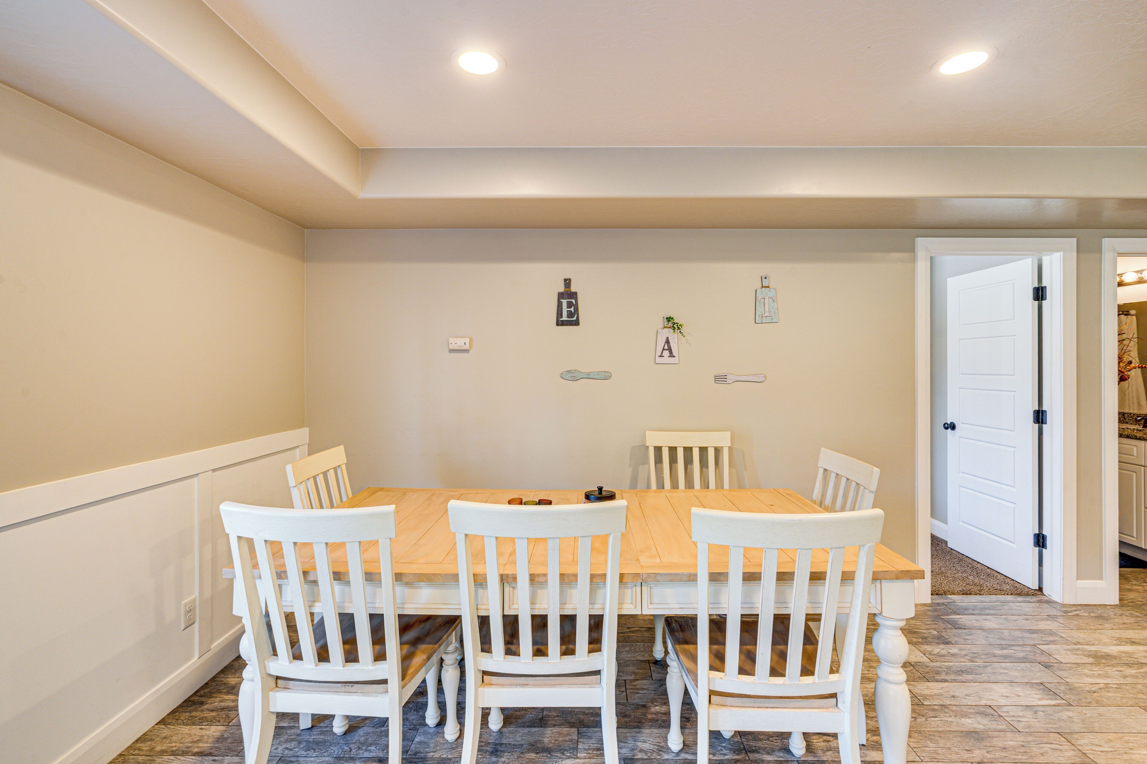 Uma sala de jantar com mesa de madeira e cadeiras brancas, luminária fixada na parede e uma porta que dá para outro cômodo.