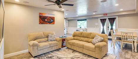 A living room with two beige sofas, a patterned rug, a ceiling fan, and a dining area with a table and chairs.