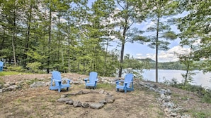 En vacker plats vid sjön med tre blå Adirondack-stolar placerade i en halvcirkel runt en eldstad, omgiven av ett naturligt landskap med träd och utsikt över sjön.