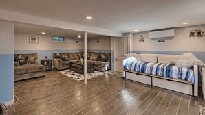 A spacious living room with a large sectional sofa, a smaller sofa, and a bed. There are multiple ceiling lights and an air conditioning unit on the wall. - Luxury Hamptons Home on Canal w/ Dock & Kayaks! (East Quogue)