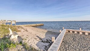 A beach with a wooden walkway leading to the water, a jetty, and buildings in the background. - Luxury Hamptons Home on Canal w/ Dock & Kayaks! (East Quogue)