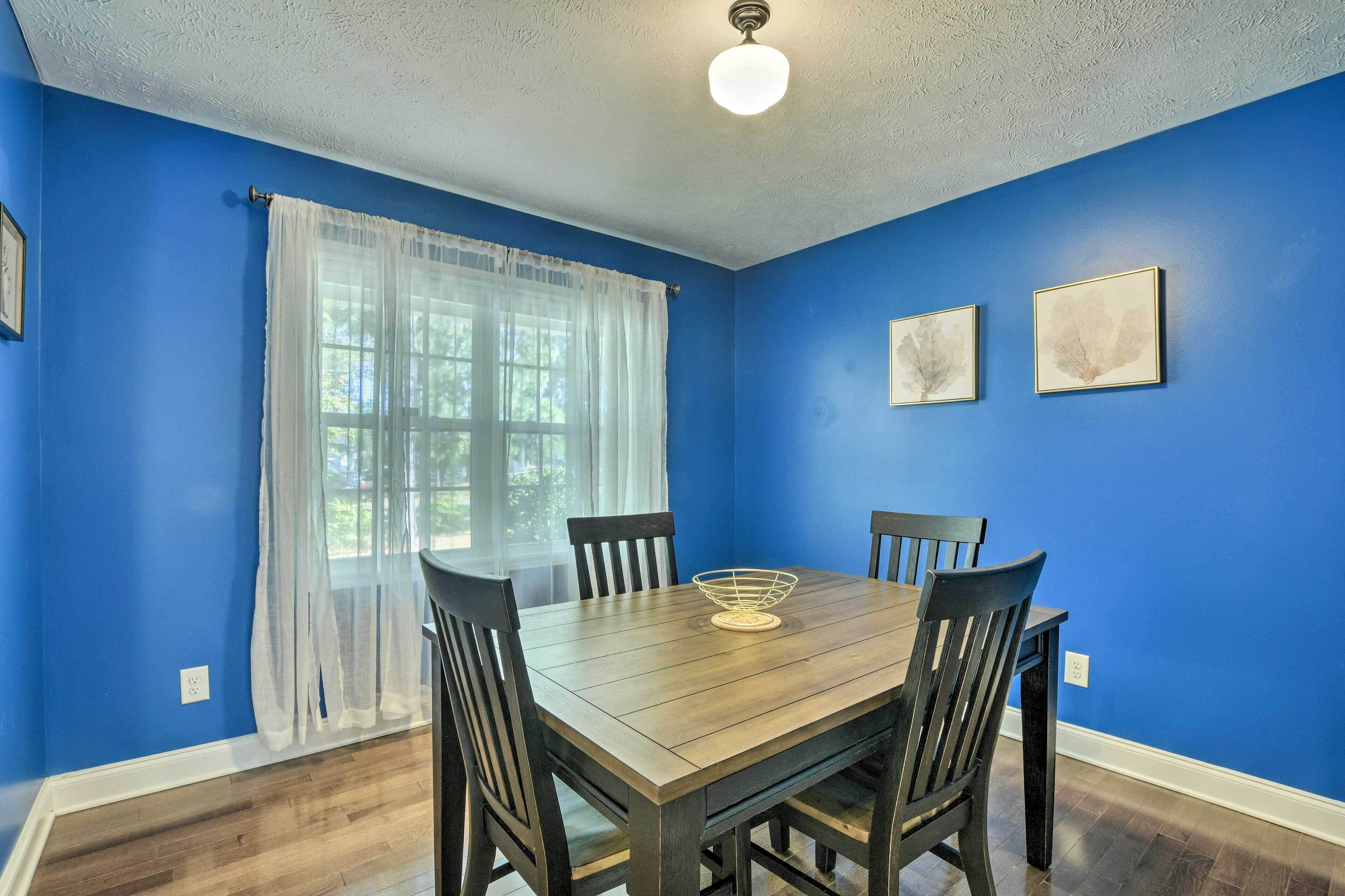 Une salle à manger avec une table et des chaises en bois, des murs bleus et une fenêtre ornée de rideaux légers.