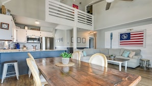 A spacious living area with a wooden dining table, a sofa, and a kitchen in the background. - Gorgeous OIB Escape w/ Dock & Canal View! (Ocean Isle Beach)