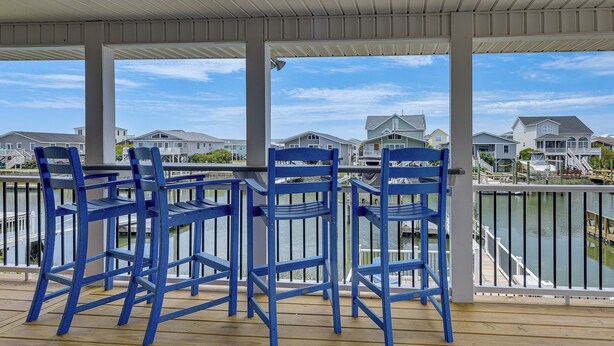 A covered outdoor area with blue chairs and a view of houses across a body of water. - Gorgeous OIB Escape w/ Dock & Canal View! (Ocean Isle Beach)