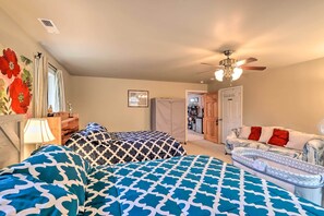 A bedroom with two beds, a ceiling fan, a window with curtains, a lamp, a framed picture, and a door leading to another room.