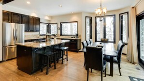 A modern kitchen with dark cabinetry, a central island, and a dining area with a chandelier. - Hot Tub & Ski Shuttle: Forested Breckenridge Home! (Breckenridge)