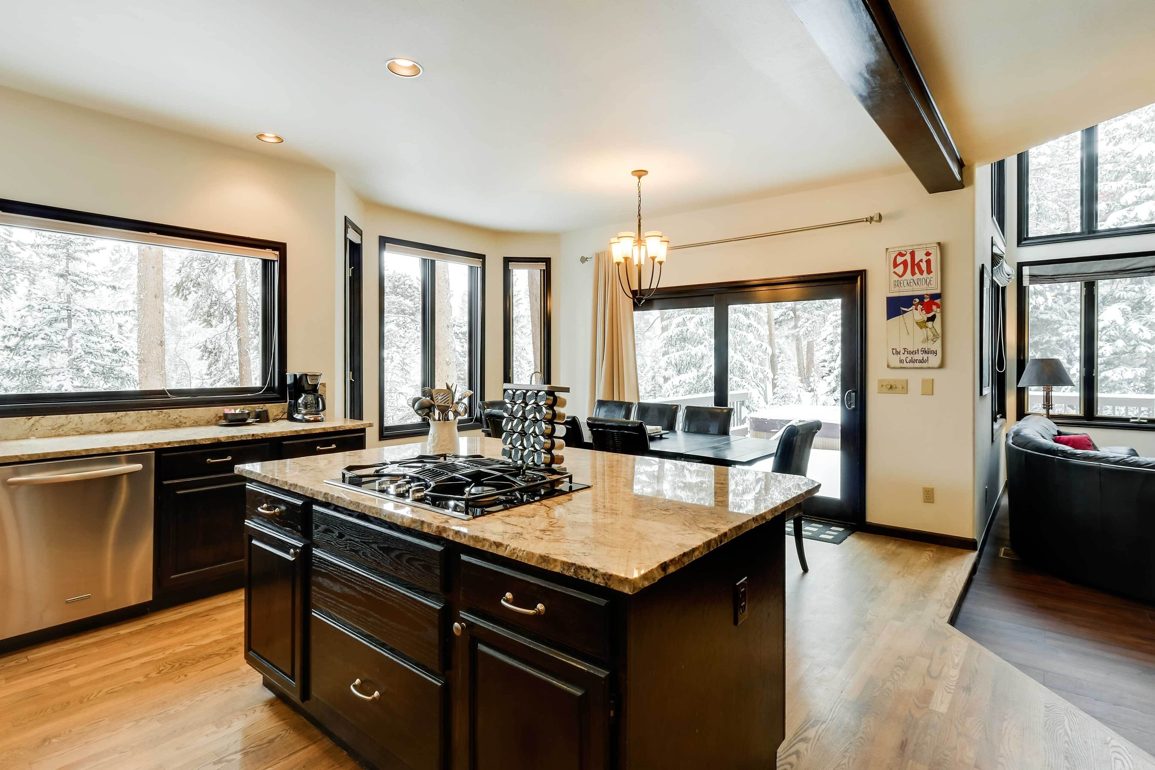 A modern kitchen with a large island, dark cabinetry, and a view of a snowy forest through large windows.