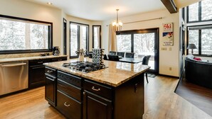 A modern kitchen with a large island, dark cabinetry, and a view of a snowy forest through large windows. - Hot Tub & Ski Shuttle: Forested Breckenridge Home! (Breckenridge)