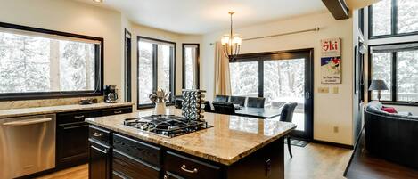 A modern kitchen with a large island, dark cabinetry, and a view of a snowy forest through large windows.