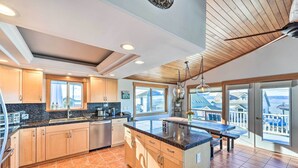A modern kitchen with wooden cabinets, a granite countertop, and a large island. There are multiple windows providing views of the surrounding area. - Mckees Beach House w/ Ocean Views, Hot Tub! (Stanwood)