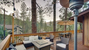 A deck with outdoor furniture, including a table and chairs, surrounded by tall trees and a view of a house. - Peaceful & Idyllic Forest Cabin w/ Pool Table (Pine Mountain Club)