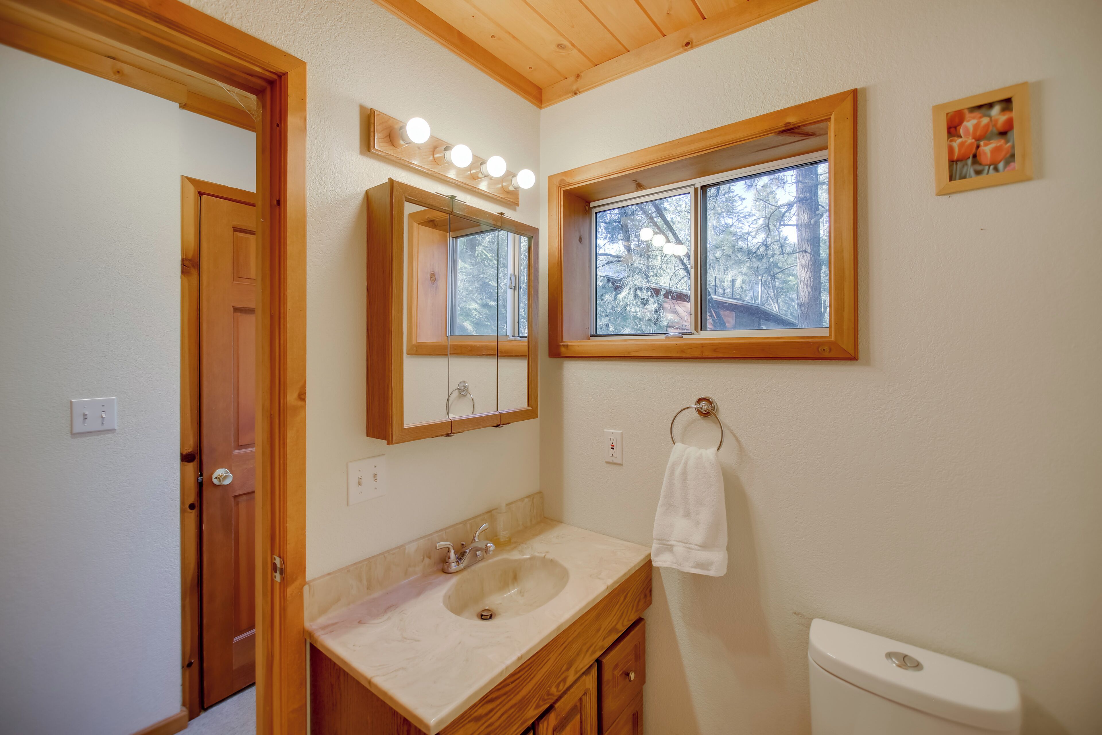 A bathroom with a wooden vanity, a sink, a mirror with lights, a window, and a toilet.