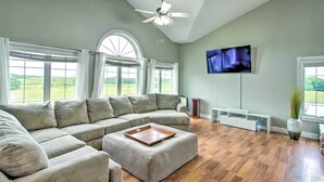 A spacious living room with a sectional sofa, a ceiling fan, a flat-screen TV, and large windows offering a view of the outdoors. - Quiet Retreat w/ Kayak & Patio, Walk to Lake! (Audubon)