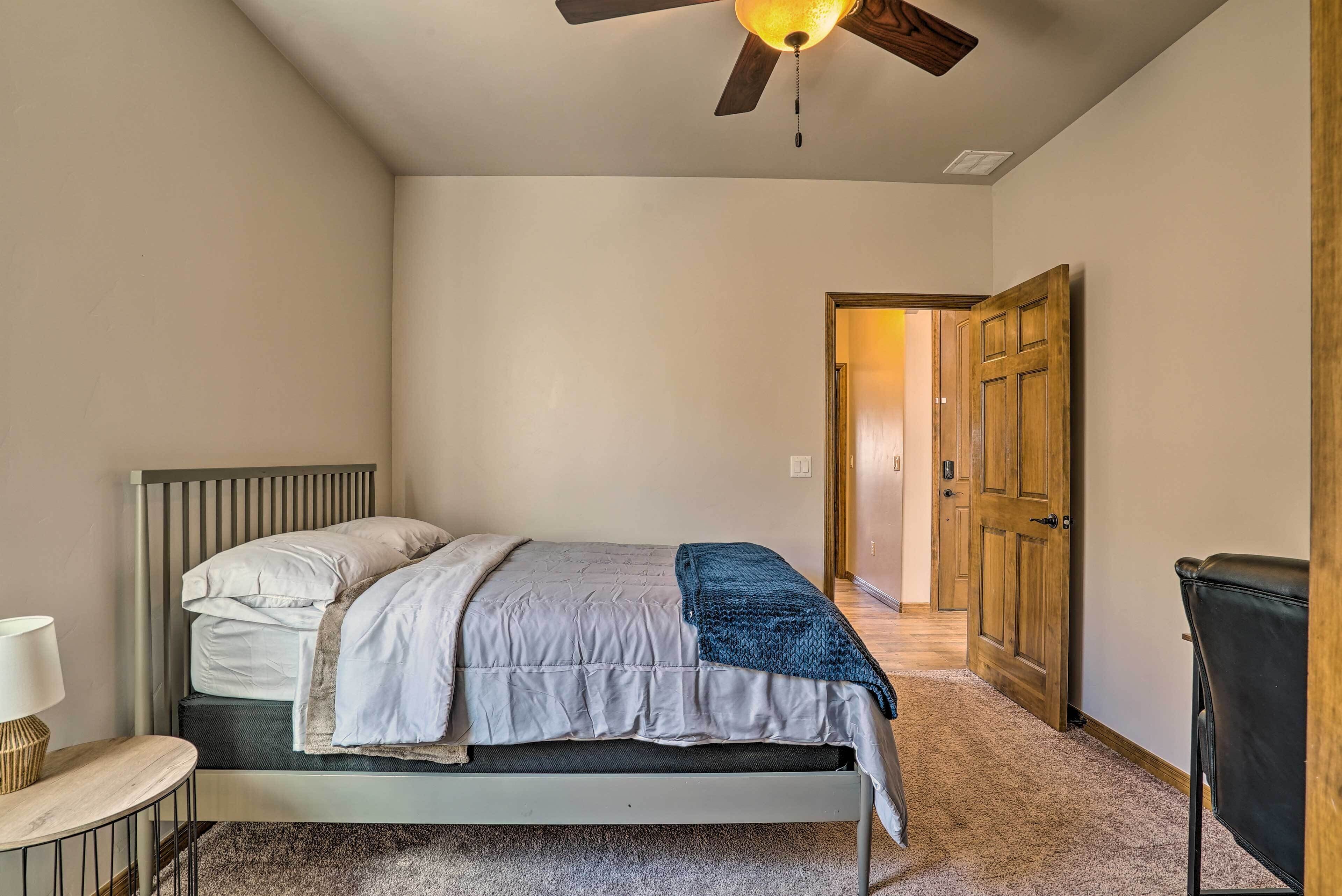 A bedroom with a bed, a bedside table, a lamp, a ceiling fan, and a door leading to another room.