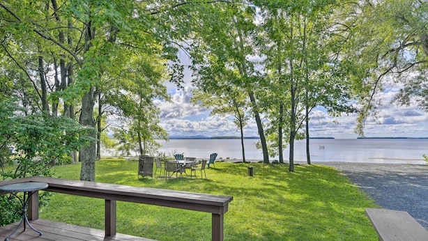 A deck with a table and chairs overlooking a grassy area with trees and a lake. - Cozy Lake Champlain Cottage w/ Private Beach! (North Hero)