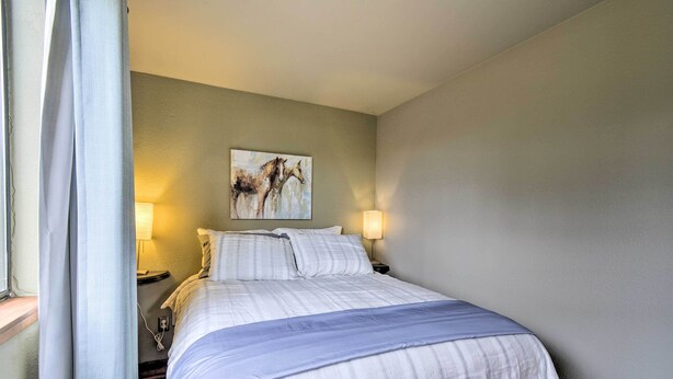 A neatly made bed with a blue blanket, two bedside lamps, a wall-mounted painting of horses, and a window with curtains.