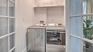 A laundry room with two front-loading washing machines, a shelf with a sign, and a window with curtains. - Indianapolis Vacation Home: 3 Mi to Dtwn! (Indianapolis)