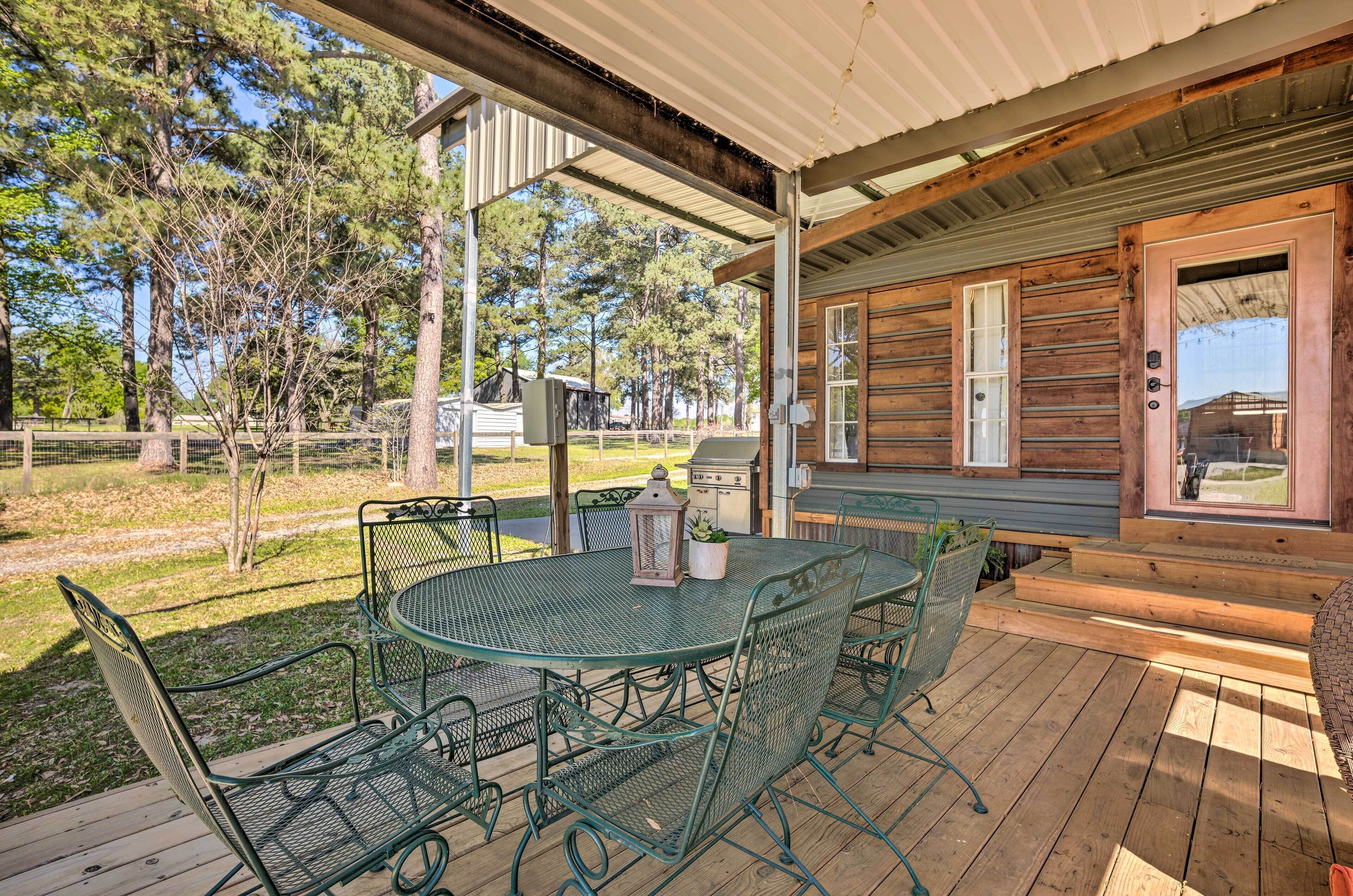 A covered outdoor patio with a round table and chairs, a grill, and a view of a wooded area.