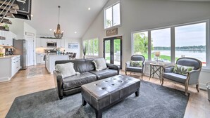 A spacious living area with a large sectional sofa, a tufted ottoman, and a dining area with a small table and chairs. There is a view of a lake through large windows. - Newly Built Gilletts Lake Gem: Dock & Hot Tub (Jackson)