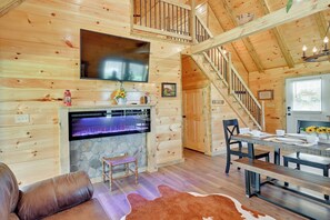 A cozy cabin interior with a stone fireplace, wooden walls, a leather sofa, a dining table with benches, and a staircase.