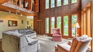 A spacious living room with a large sectional sofa, a pink armchair, and a chaise lounge. The room features wooden walls and large windows offering views of the outdoors. - Waupaca Lakefront Home w/ Pool Table & Dock! (Waupaca)