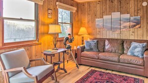 A cozy cabin interior with a wooden paneled wall, a leather sofa, a wooden chair, and a tripod-mounted camera.
