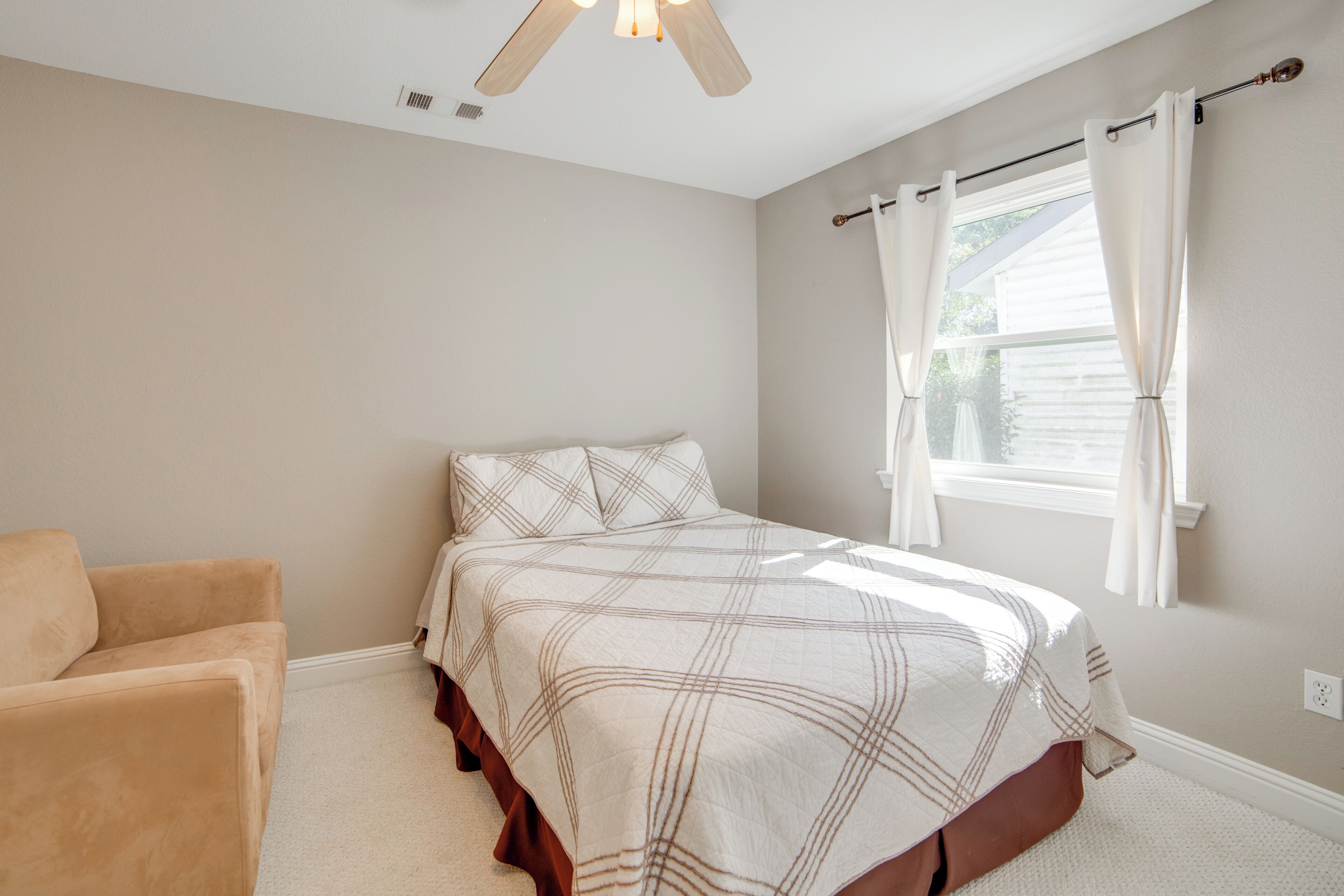 A bedroom with a bed, a chair, a window with curtains, and a ceiling fan.
