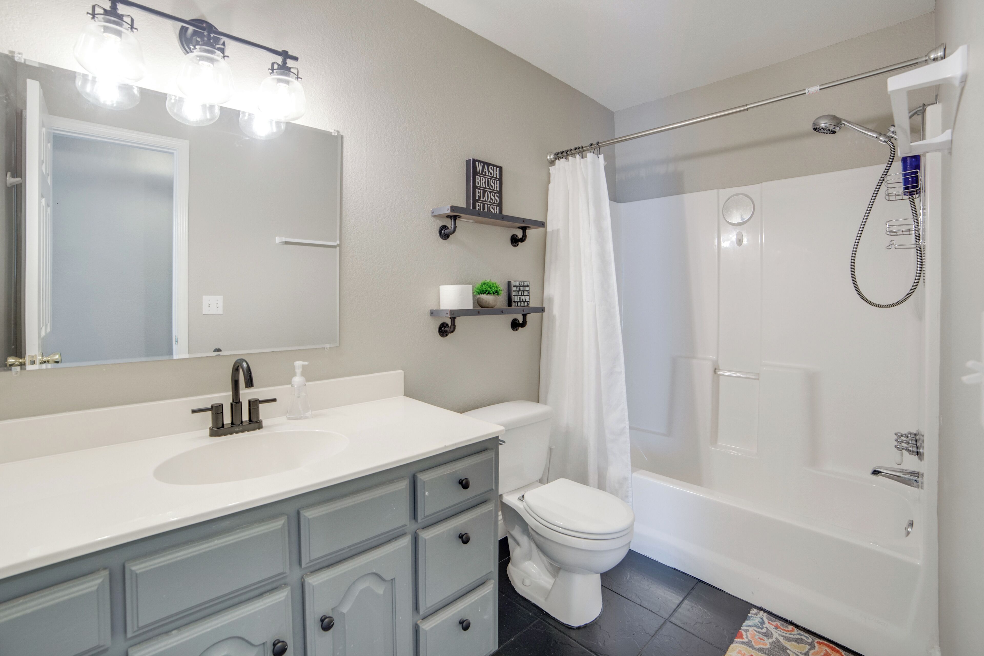 Een moderne badkamer met een witte wastafel, meubel en toilet. Boven de wastafel zijn er drie planken, waarvan er één een potplant en een bordje bevat, en een andere een klein vaasje. Een douchegordijn is zichtbaar, en een handdouche is aan de muur bevestigd.