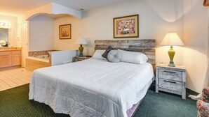 Une chambre à coucher bien aménagée, avec un grand lit, une tête de lit en bois, deux tables de chevet avec des lampes, une petite salle de bain et un tapis à motifs verts.