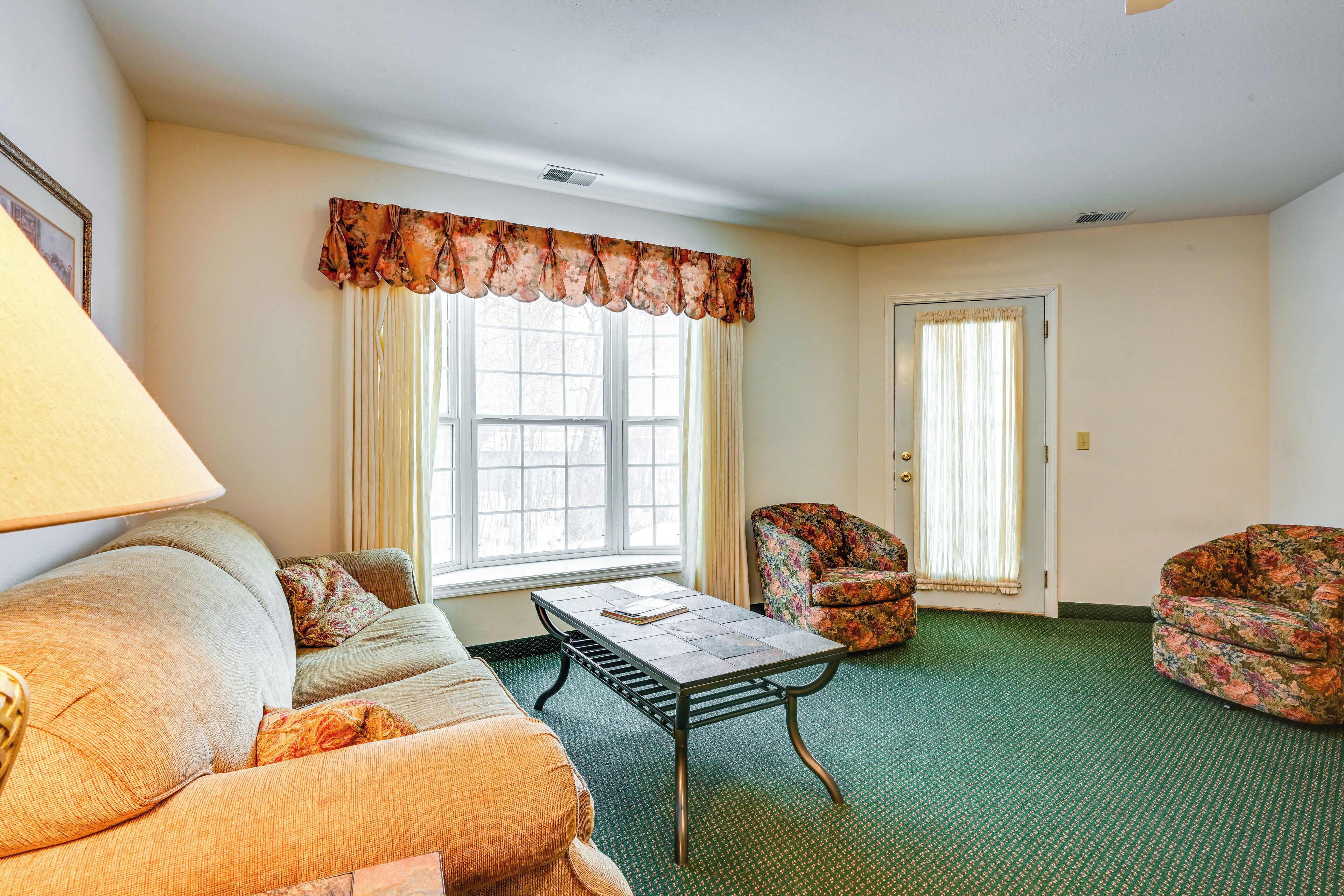 A living room with a sofa, a coffee table, and two floral armchairs. There is a window with curtains and a door in the background.