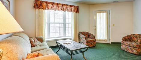 A living room with a sofa, a coffee table, and two floral armchairs. There is a window with curtains and a door in the background.