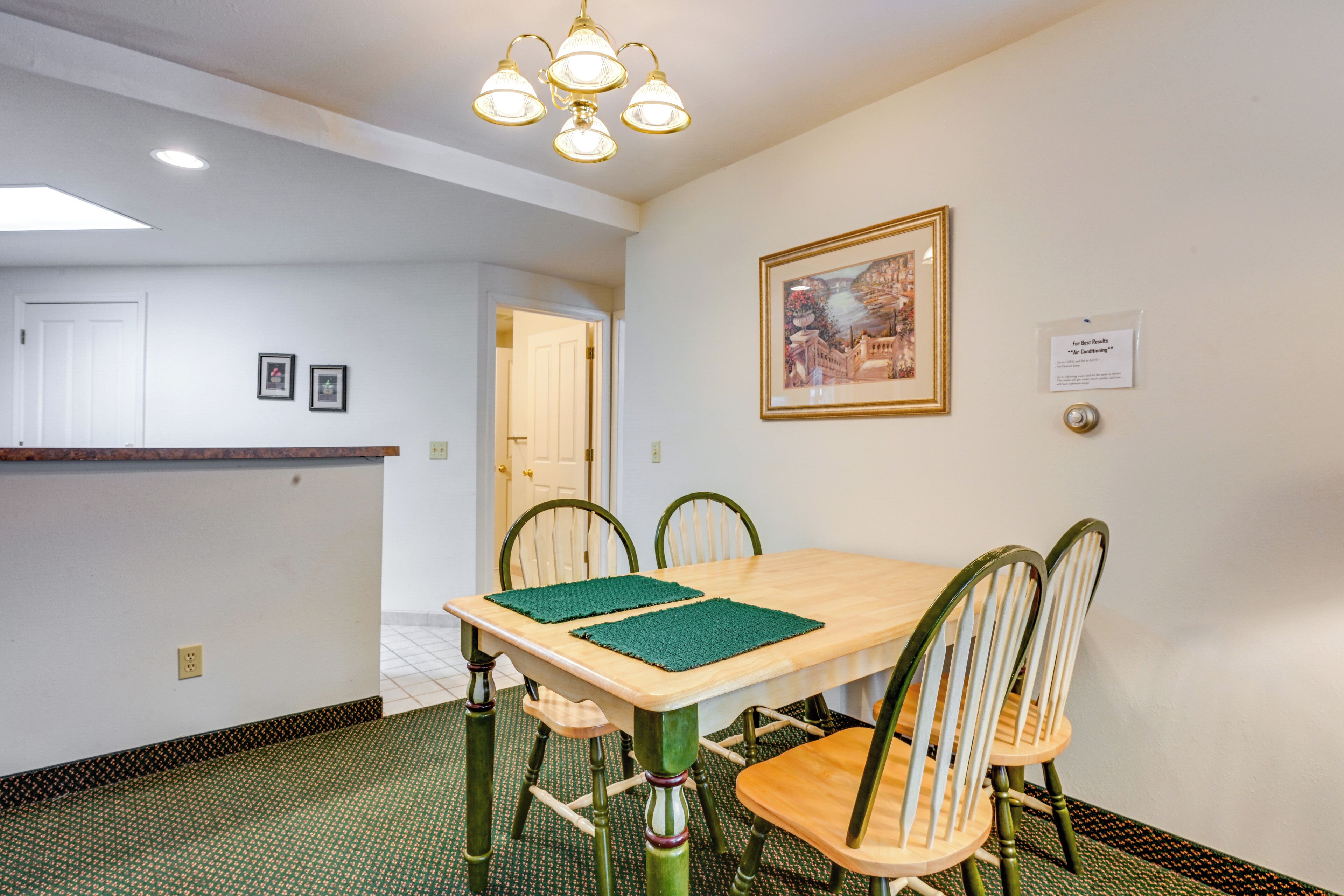 A dining area with a wooden table, four chairs, a green placemat, a framed picture on the wall, and a door in the background.