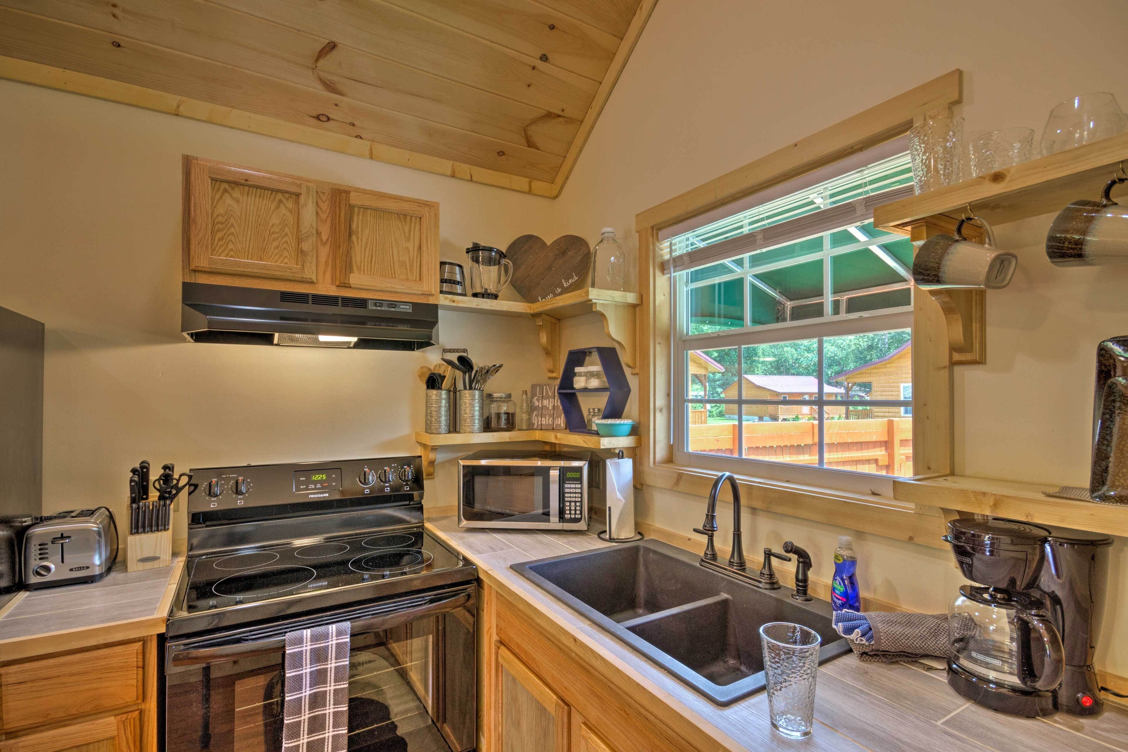 A compact kitchen with a stove, microwave, sink, and coffee maker. There are wooden cabinets, a window with a view, and various kitchen utensils.