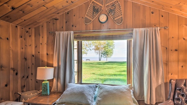 Un dormitorio de cabaña de madera con una cama, una mesita de noche, una lámpara y vistas a un campo cubierto de hierba.