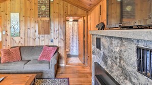 Interior acogedor de una cabaña con techo de madera, chimenea de piedra, sofá con cojines y televisor sobre una pared de piedra.