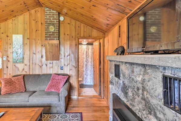 L’intérieur chaleureux d’un chalet avec un plafond en bois, une cheminée en pierre, un canapé avec des coussins et un téléviseur fixé à un mur en pierre.