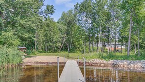 A wooden dock extending into a calm body of water, surrounded by dense green trees and a sandy area with a small structure in the background.