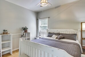 A bedroom with a white bed, a bedside table, a nightstand, a window with curtains, and a ceiling fan.