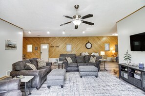 A modern living room with a ceiling fan, a large sofa set, a coffee table, and a TV mounted on the wall.