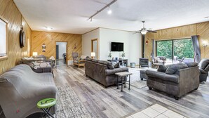 A spacious living room with a large sofa, a smaller couch, and a coffee table. There is a television mounted on the wall, and a window providing natural light.