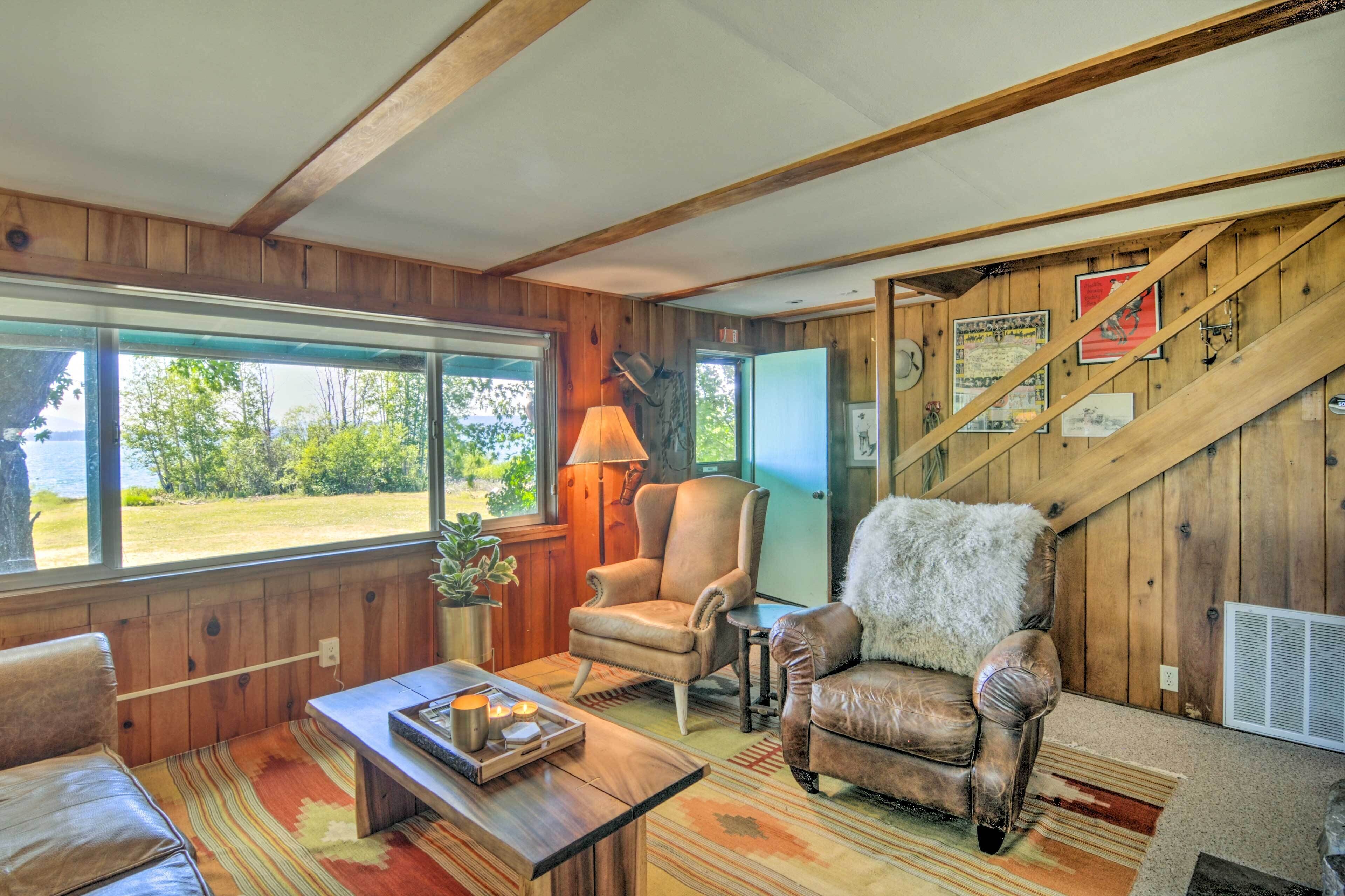 A cozy cabin interior with a wooden interior, a large window offering a view of the outdoors, a leather armchair, a small table with a tray, and a lamp.