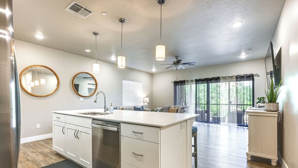 A modern kitchen with white cabinetry, a dishwasher, and a large island with a sink. There are pendant lights, a ceiling fan, and a view of a living area with a couch and a plant. - Resort Pool & Hot Tub: Modern Condo in St George! (St. George)