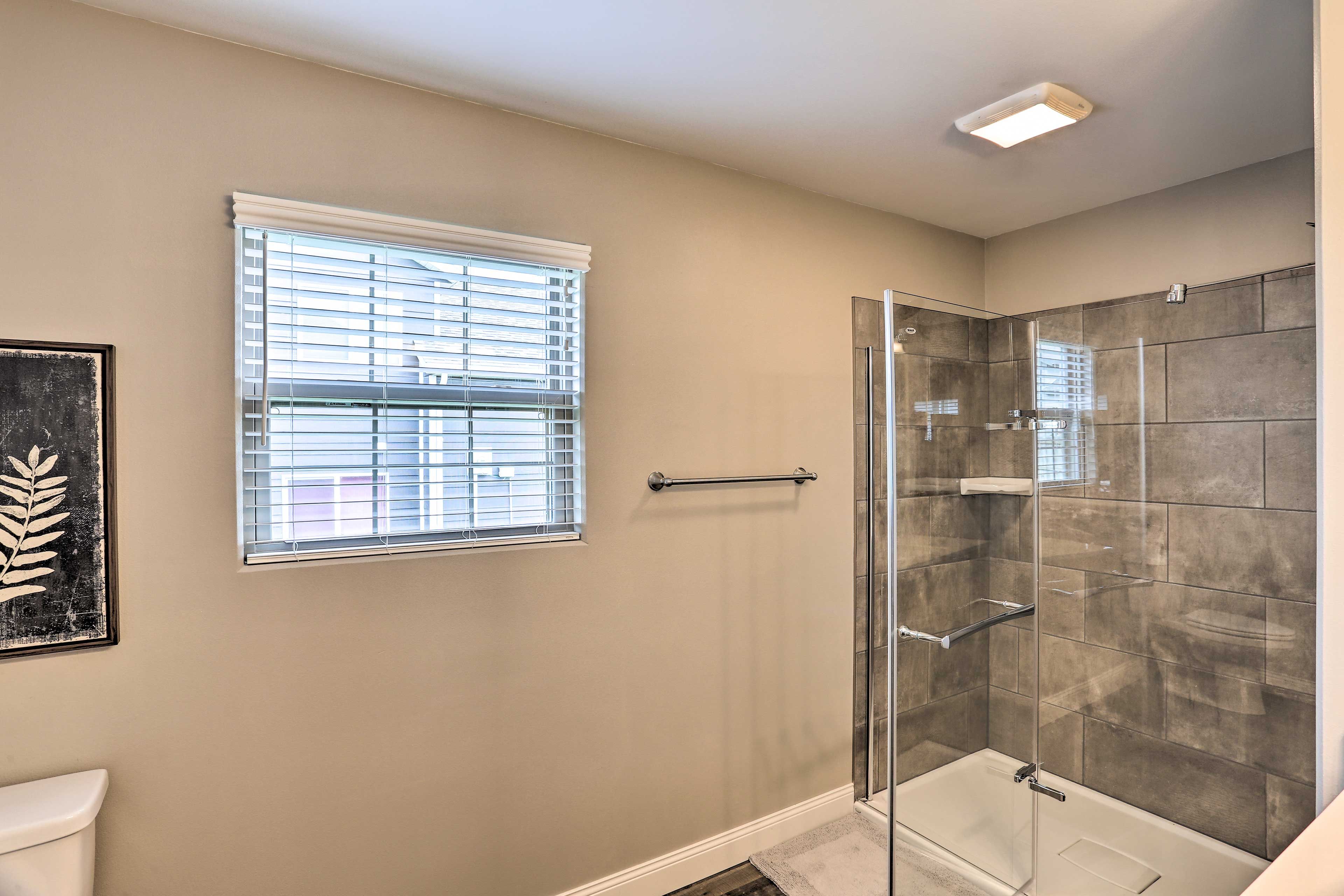 A bathroom with a glass-enclosed shower, a toilet, and a window with blinds.