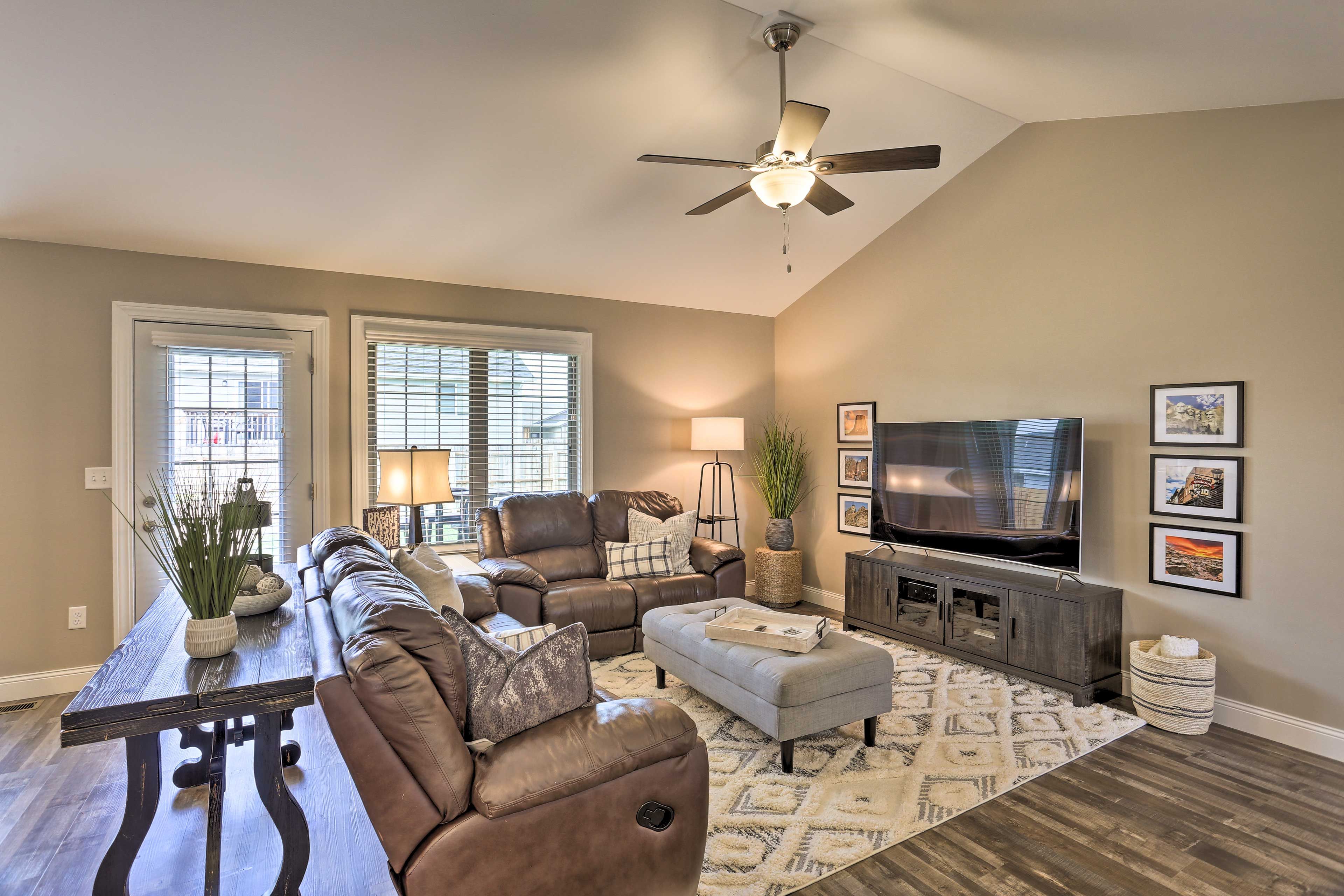 A spacious living room with a large sofa, a coffee table, and a television. There is a ceiling fan and a window with blinds.