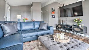 A modern living room with a blue leather sofa, a grey tufted ottoman, a flat-screen TV, and a console table with decorative items.