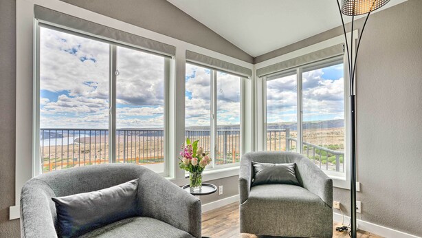 A room with two grey armchairs, a small table with a vase of flowers, and large windows offering a view of a fenced area and distant landscape.
