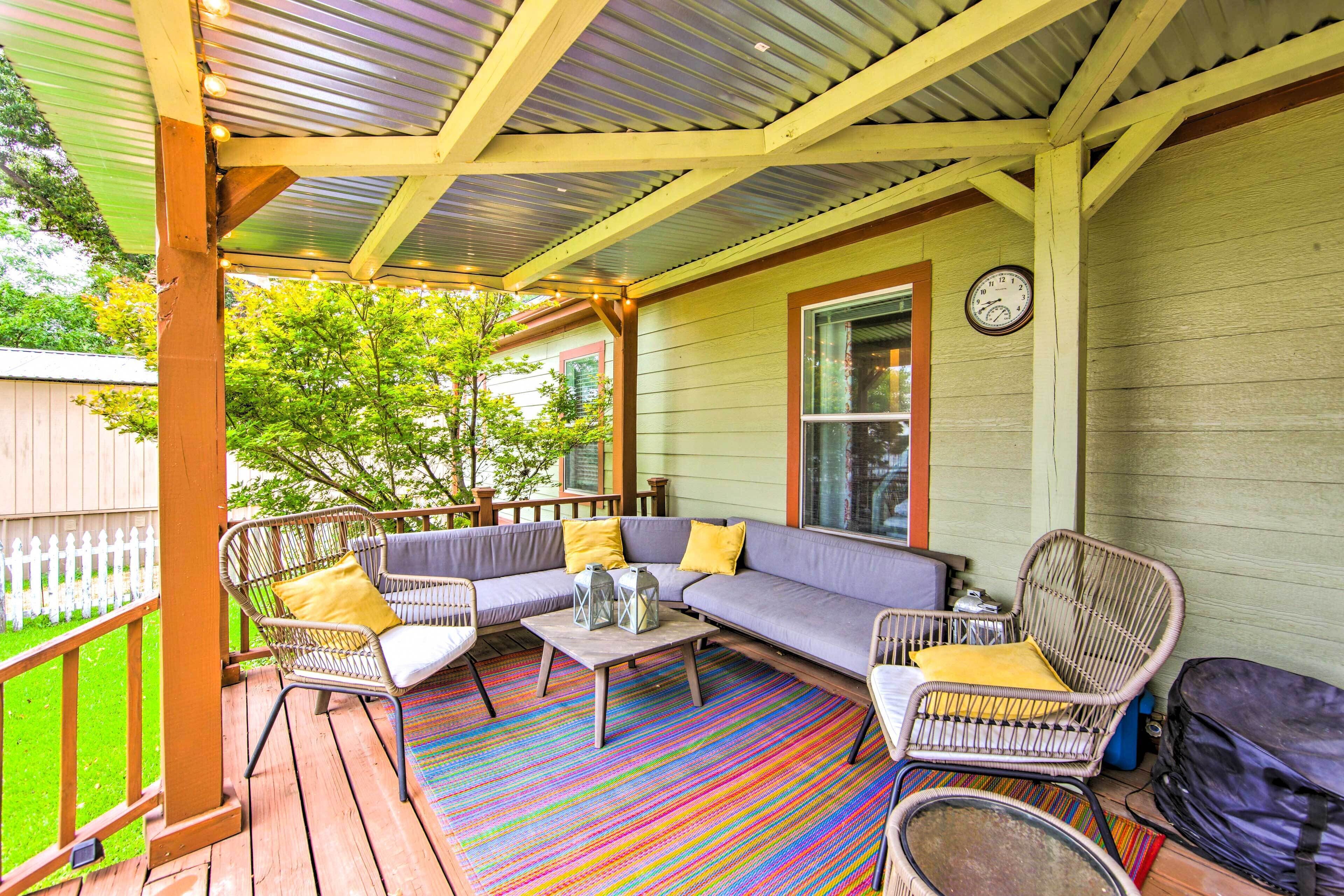 A covered porch with a colorful striped rug, a gray sofa with yellow cushions, wicker chairs, and a clock on the wall.