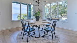 A dining area with a round table, six chairs, a central vase with greenery, and large windows offering a view of the sea. - Entire Luxury Lake Granbury Cliffside Home w/ Deck (Granbury)