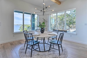 A dining area with a round table, six chairs, a central vase with greenery, and large windows offering a view of the sea. - Entire Luxury Lake Granbury Cliffside Home w/ Deck (Granbury)