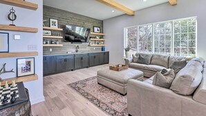 A modern living room with a beige sofa, a wooden ceiling, a large window, and a kitchen area with a TV and shelves.